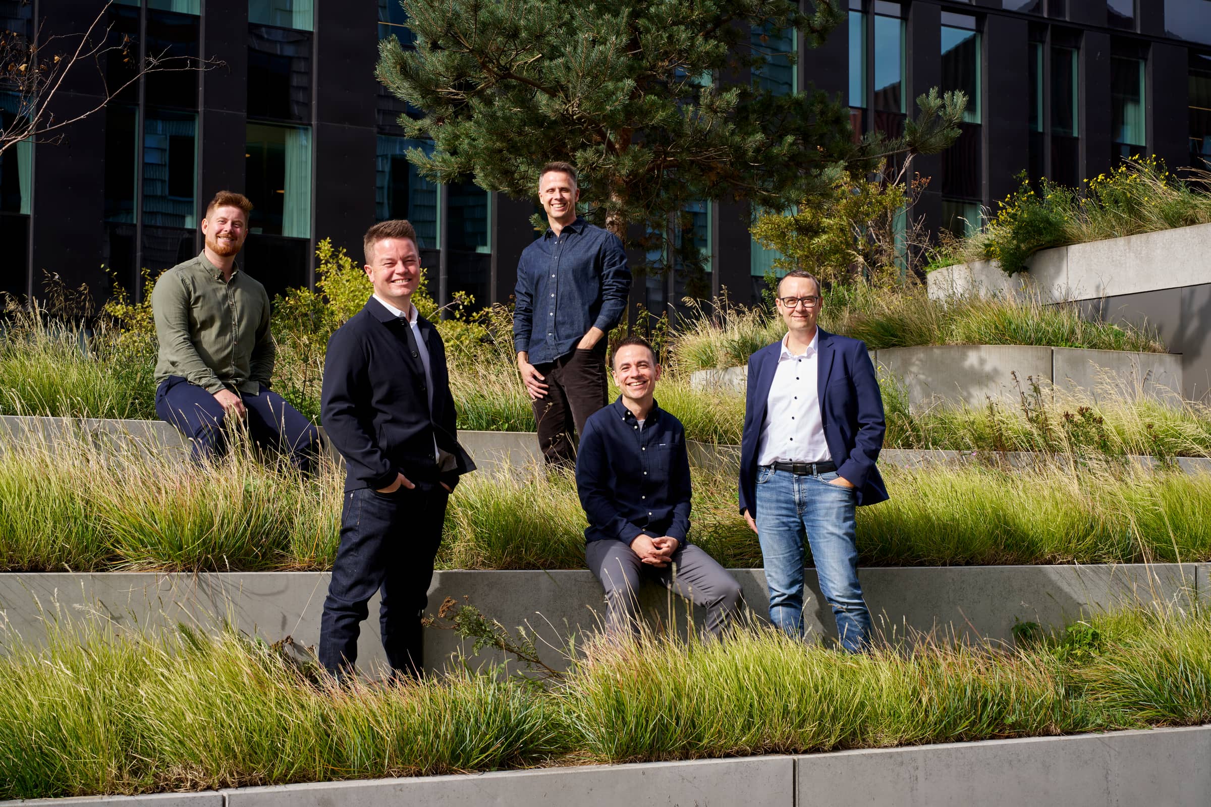 The five Distinkt partners outdoors on terraced concrete planters full of tall grass, in front of a dark office building and a pine tree, smiling at the camera.